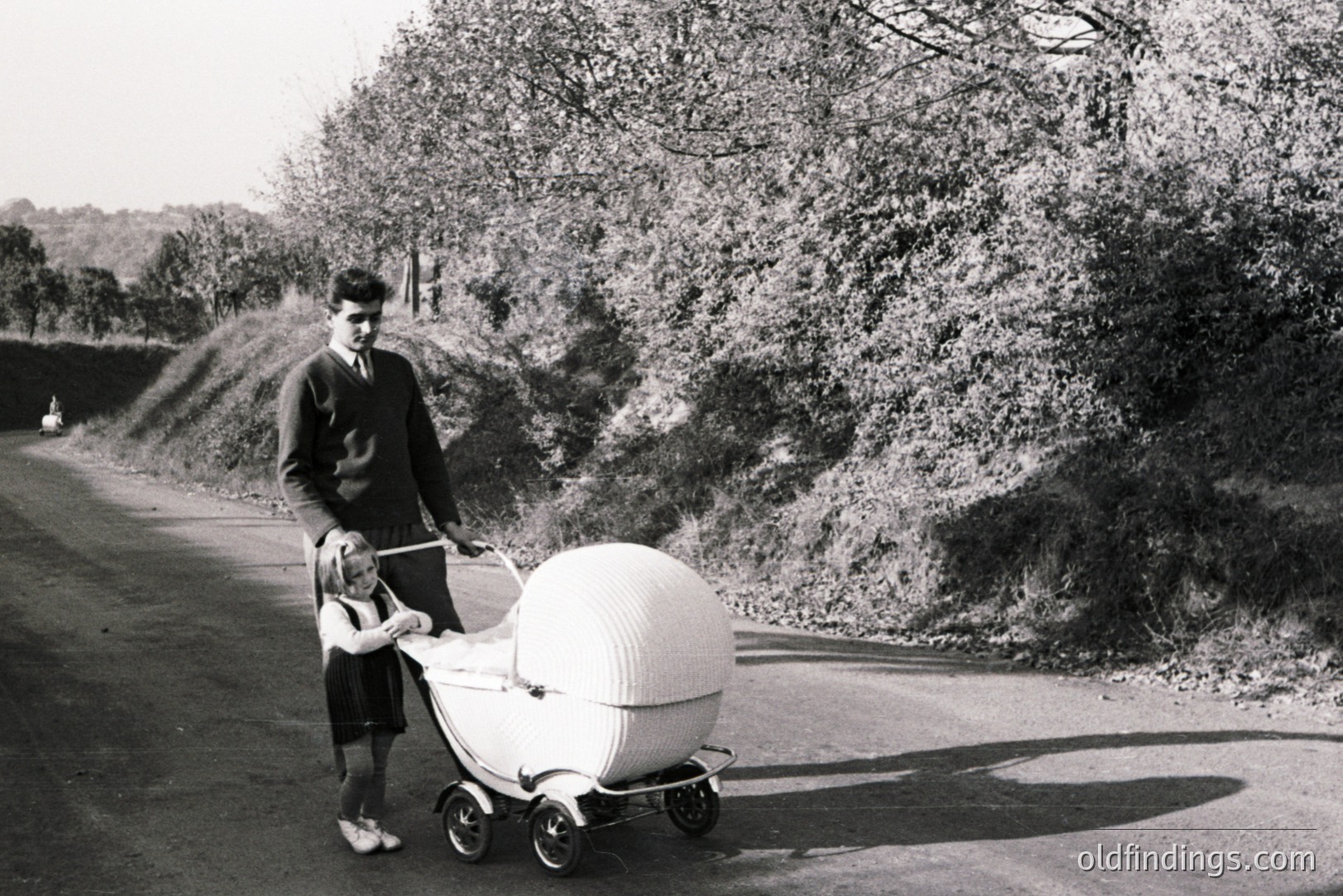 A young man in a suit and tie guides a child with a unique, rounded baby carriage along a tree-lined road. The carriage's design suggests mid-century modern influence. Likely 1960s/1970s, rural setting. A sense of domesticity and nostalgic style. Could be of interest for design reference.