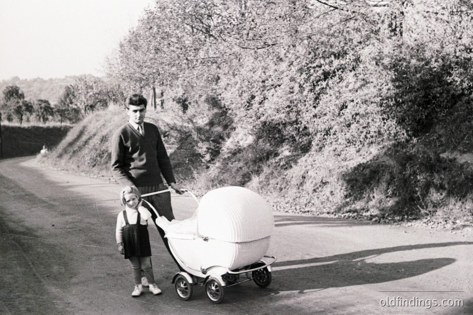 Man in a sweater and tie stands with a young girl beside an unusual, futuristic-looking pram on a rural road. Likely mid-20th century, perhaps 1960s. A striking example of modernist design intersecting with family life. A visually compelling image, potential for design reference.
