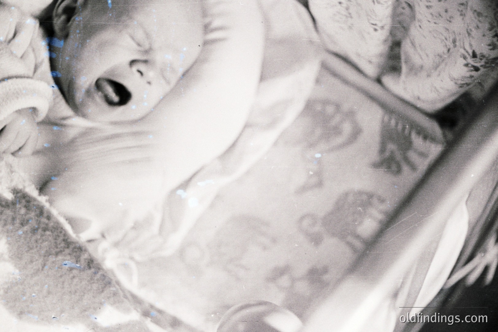 A baby in a crib, captured in a candid moment of distress or surprise. The child's mouth is open in a cry or gasp. Crib railing visible. Vintage photograph shows age with surface markings and faded tones. Likely mid-20th century family snapshot. Subject matter appealing for nostalgia and family history collections.