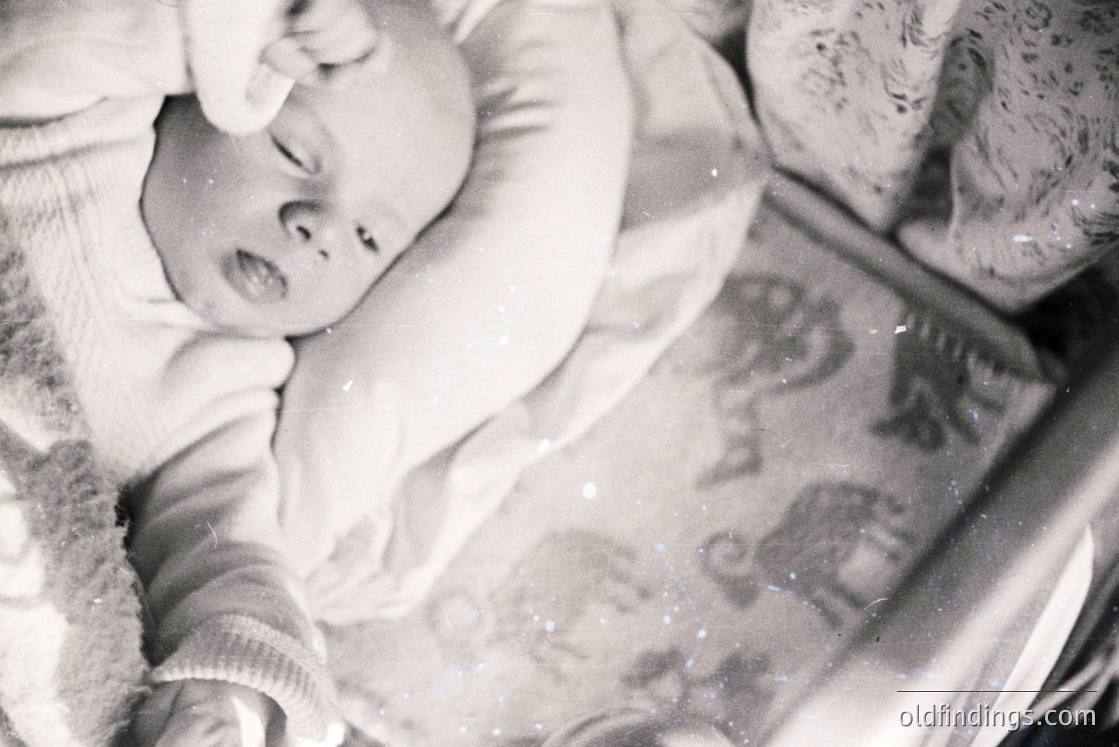 A baby sleeps peacefully in a crib covered with a patterned blanket featuring animal illustrations. The monochrome image shows wear consistent with age and usage. Likely a family snapshot, c. 1960s-1970s. Captures a moment of childhood innocence.