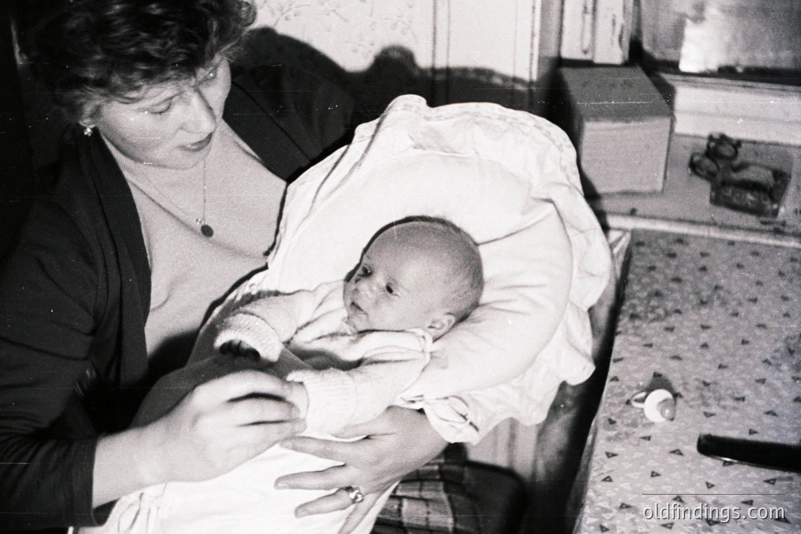 A young woman cradles a newborn baby wrapped in a blanket. Likely a domestic interior scene, with visible patterned flooring and simple furnishings. Appears to be a snapshot, possibly from the 1960s-1970s. Jewelry and clothing suggest a modest lifestyle. Nostalgic family moment captured.