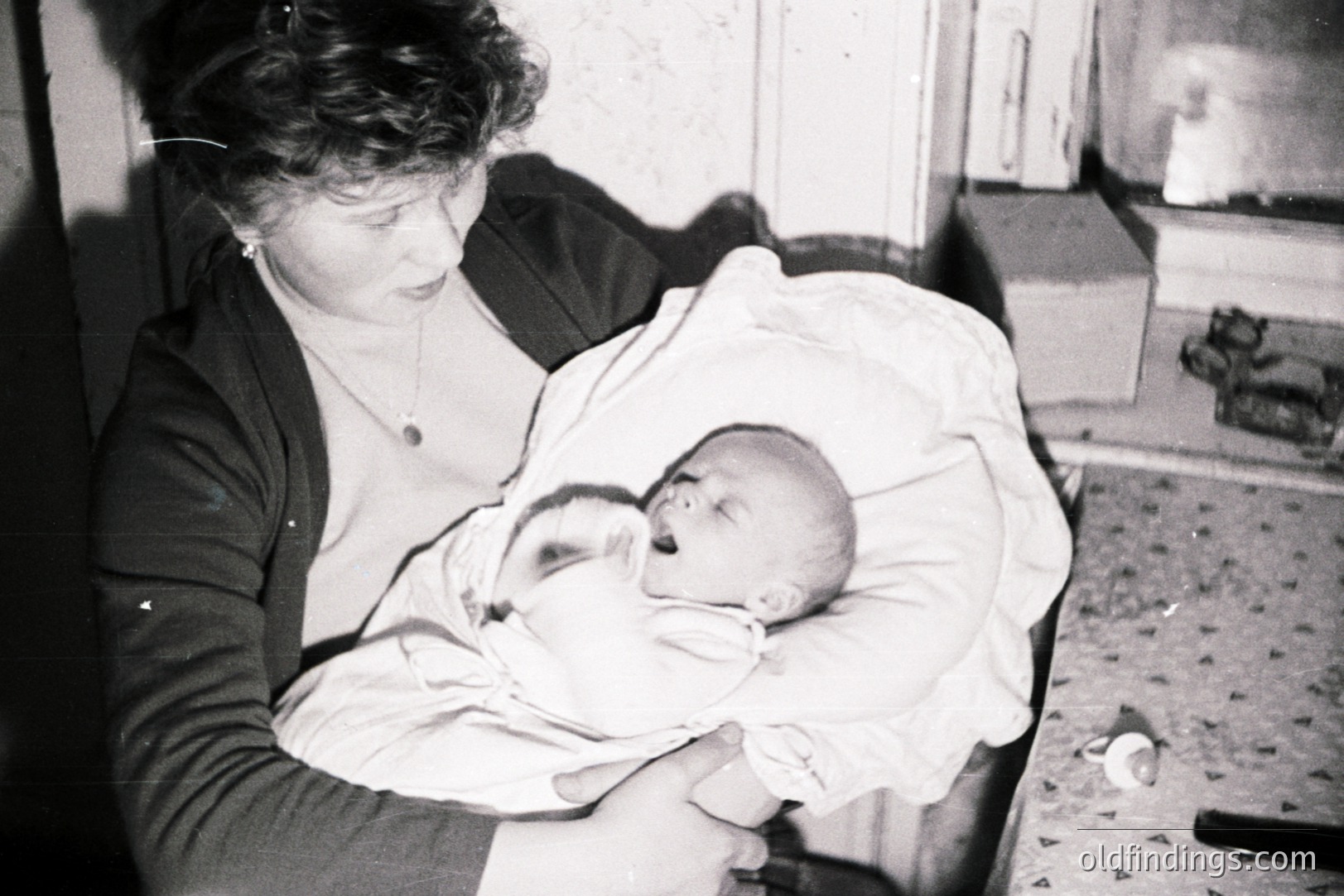 A woman holds a swaddled infant in her arms, viewed from above. Interior setting; likely a domestic space with a tiled floor and simple furnishings. The woman wears a dark dress & necklace. Possible 1950s-1960s style. Subject matter suitable for family history research or vintage portrait design.