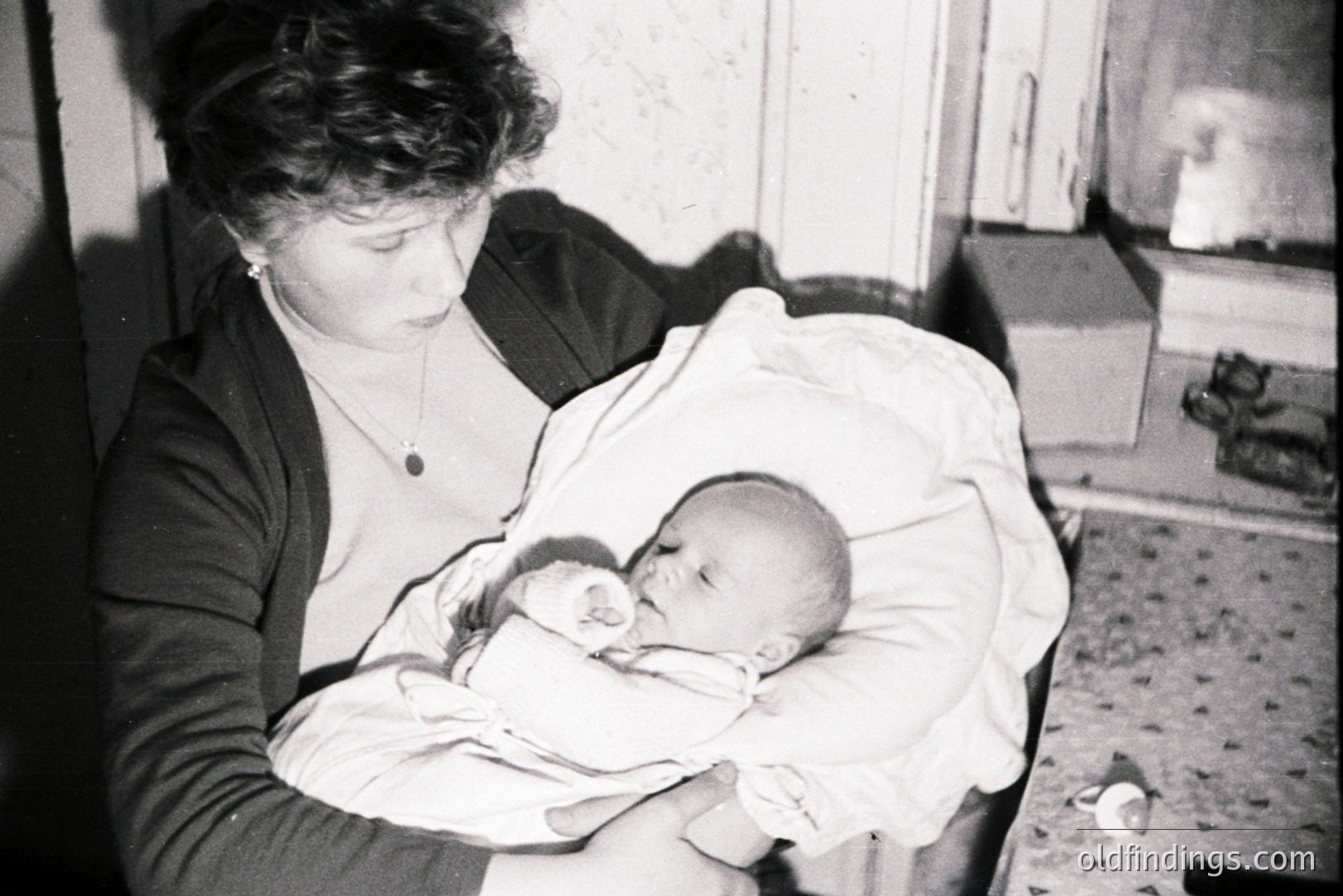 A young woman holds a swaddled infant close, viewed in a dimly-lit, interior setting. She wears a dark cardigan and a simple necklace. The infant appears asleep. The backdrop includes simple furniture and textured walls. Likely a domestic snapshot, c. 1950s-1970s.