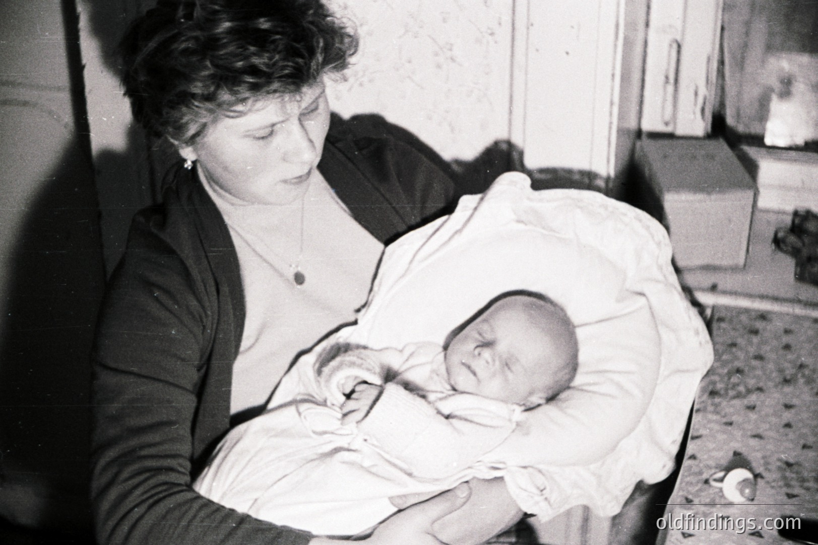 A mother tenderly cradles a sleeping baby wrapped in a light-colored blanket. The scene is intimate, set within a dimly lit interior with a visible door and simple furnishings. Likely a family portrait from the mid-20th century. The mother wears a cardigan and a necklace. This image holds sentimental and historical research value.