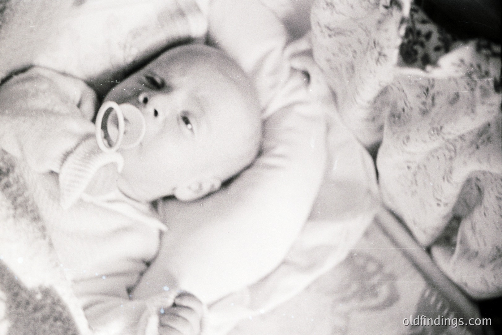Close-up black and white photograph of an infant, likely a baby, bundled in a textured blanket. The child holds a pacifier in their mouth. Soft lighting and shallow depth of field indicate a personal, intimate moment. Appears to be a snapshot from the 1950s-1970s.