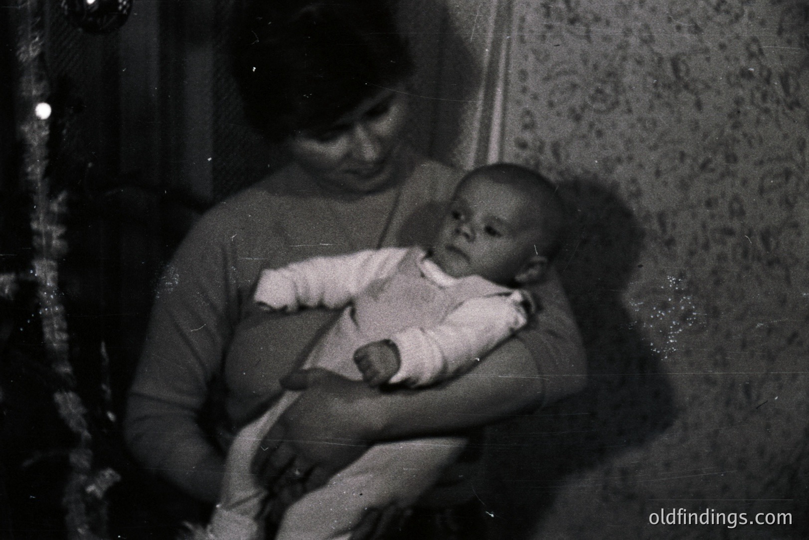 A mother cradles an infant in her arms, set against a textured wallpaper backdrop and hinting at a Christmas tree. The image, captured in black and white, conveys a candid, intimate family moment. Likely a snapshot from the 1960s or 70s. Represents domestic life and interpersonal connection.