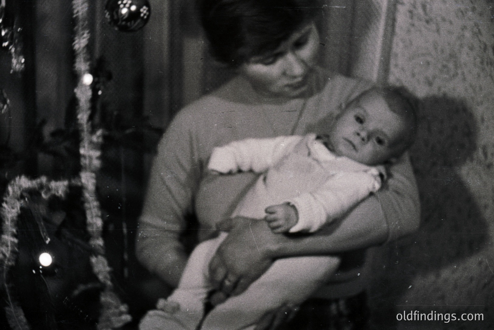 Black and white image depicts a woman holding a baby. The woman wears a turtleneck sweater; the infant is in a white onesie. A Christmas tree and garland are visible in the blurred background, suggesting a holiday setting. Likely 1960s-1970s, family portrait style.