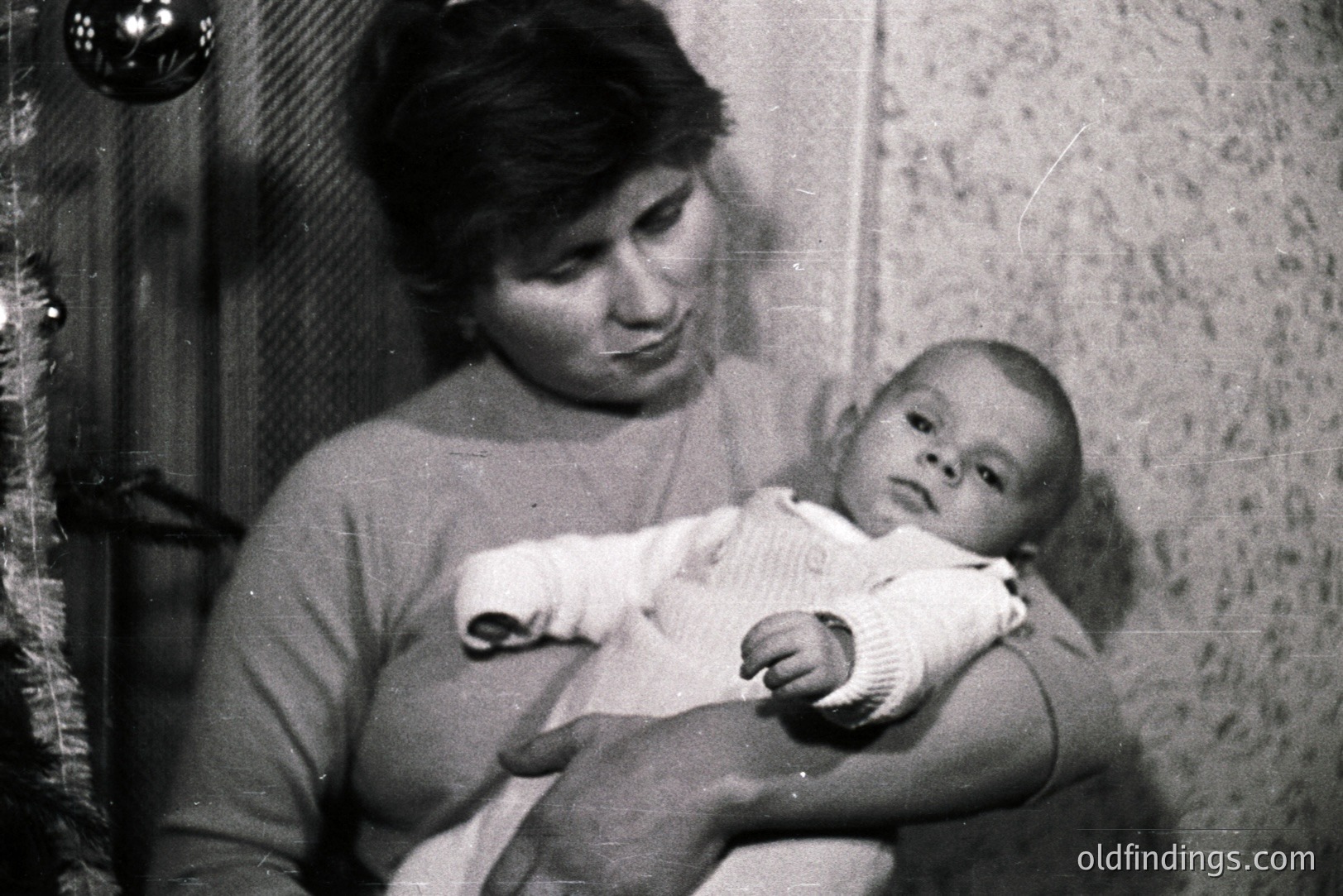 A mother cradles her infant, likely indoors. The scene features a textured wall and a partial view of a Christmas tree with ornaments. The image suggests a domestic moment, possibly a family portrait, c. 1960s-1970s. Commercial value: sentimental family studies.