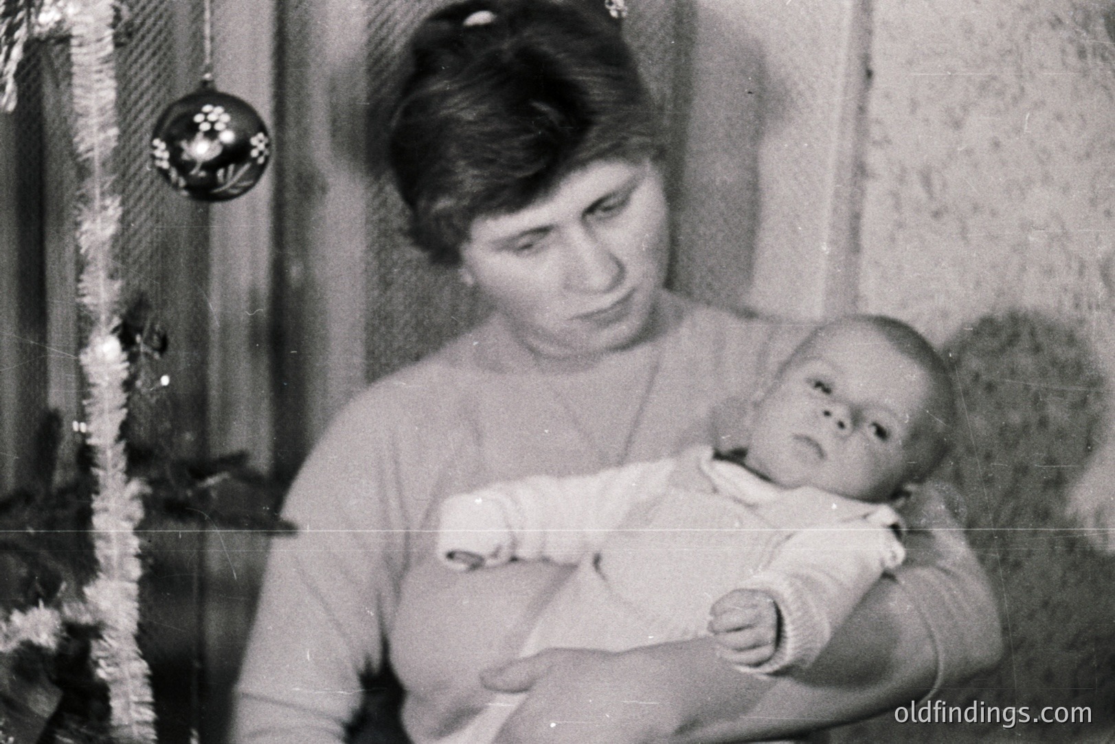 A young woman cradles a baby in her arms, posing near a Christmas tree decorated with a single ornament. A gentle, intimate portrait, likely a family snapshot. The image’s grainy quality and style suggest a 1960s or 1970s origin. Subject likely Eastern European based on facial features.