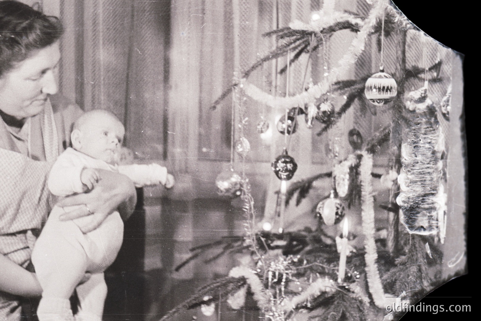 A mother gently holds a baby in front of a decorated Christmas tree. The tree features tinsel, glass ornaments, and traditional candles. Likely a domestic scene from the mid-20th century, showing festive family life. Shows wear and tear consistent with an older print.