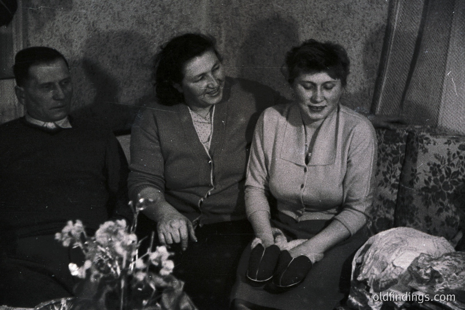 A formal portrait depicts a man and two women seated, appearing slightly melancholic. Floral wallpaper and an upholstered chair suggest an interior setting. The women wear cardigans and dresses, typical of the 1960s-1970s. A pair of shoes sits in the foreground, adding an intimate, domestic detail.