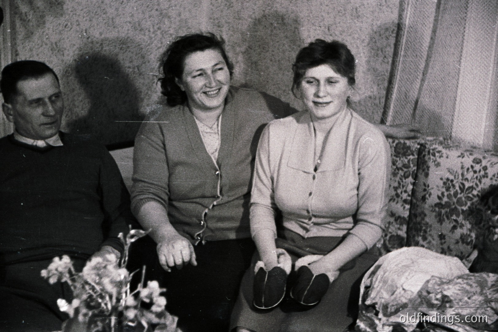 A black and white snapshot of a man, woman, and young woman seated on a floral patterned sofa. The man wears a dark sweater. The women are in cardigans, smiling slightly. A child is partially visible at right. Likely a family portrait, circa 1960s-1970s. Interior scene with visible window and curtain.