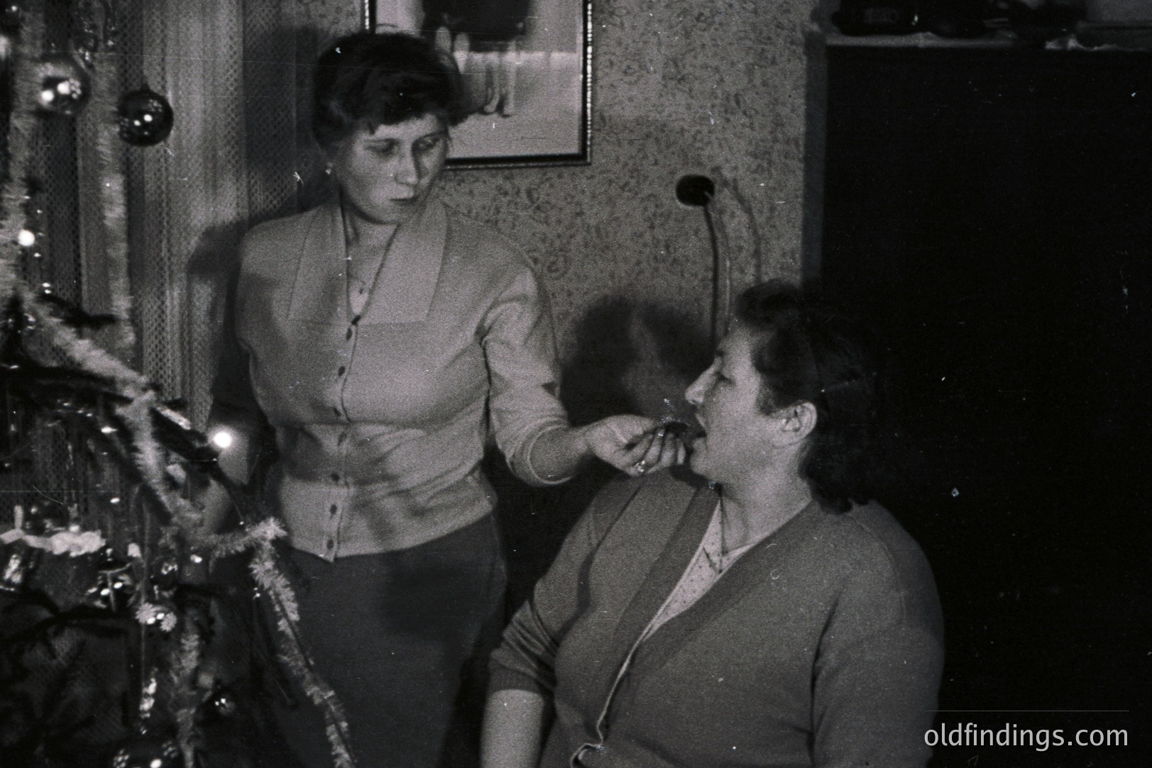 Two women near a decorated Christmas tree. One offers the other a cigarette. Interior scene with textured wallpaper & traditional décor. Likely a domestic moment, possibly mid-1950s or early 1960s. Captures social customs of the era.
