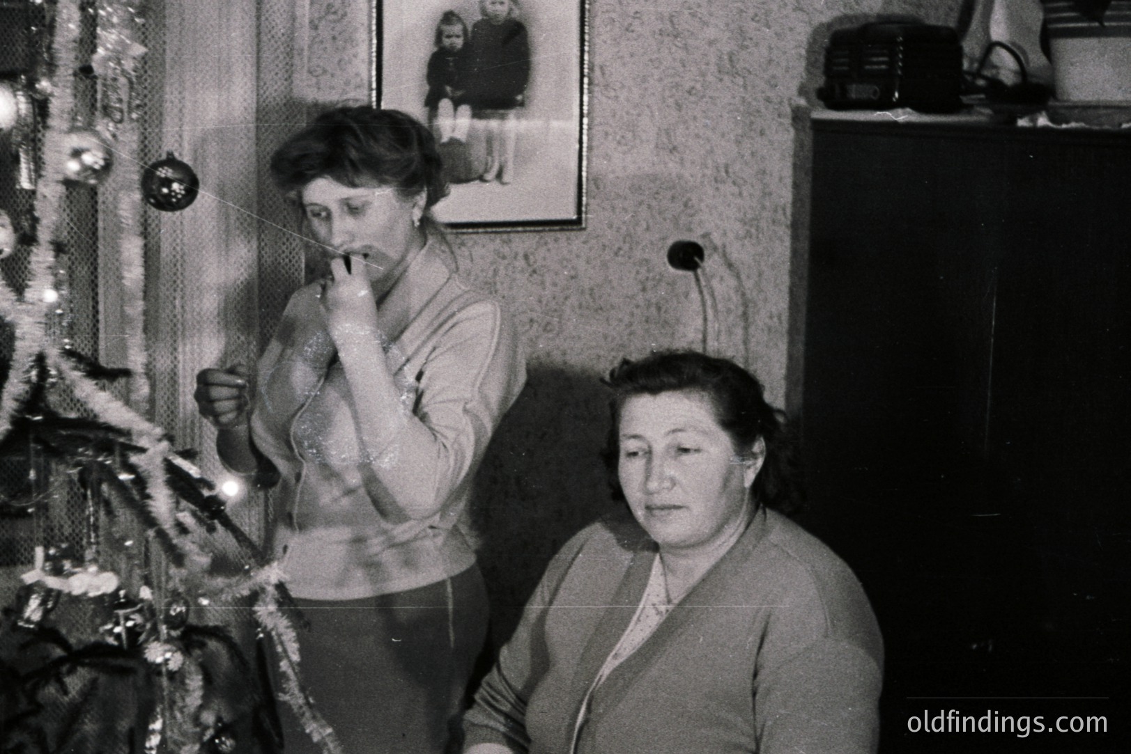 Black and white photo depicting two women indoors, one decorating a Christmas tree, the other seated, seemingly observing. Mid-century modern decor evident; framed photo on wall. Likely a family snapshot, capturing domestic life. Estimated 1960s.