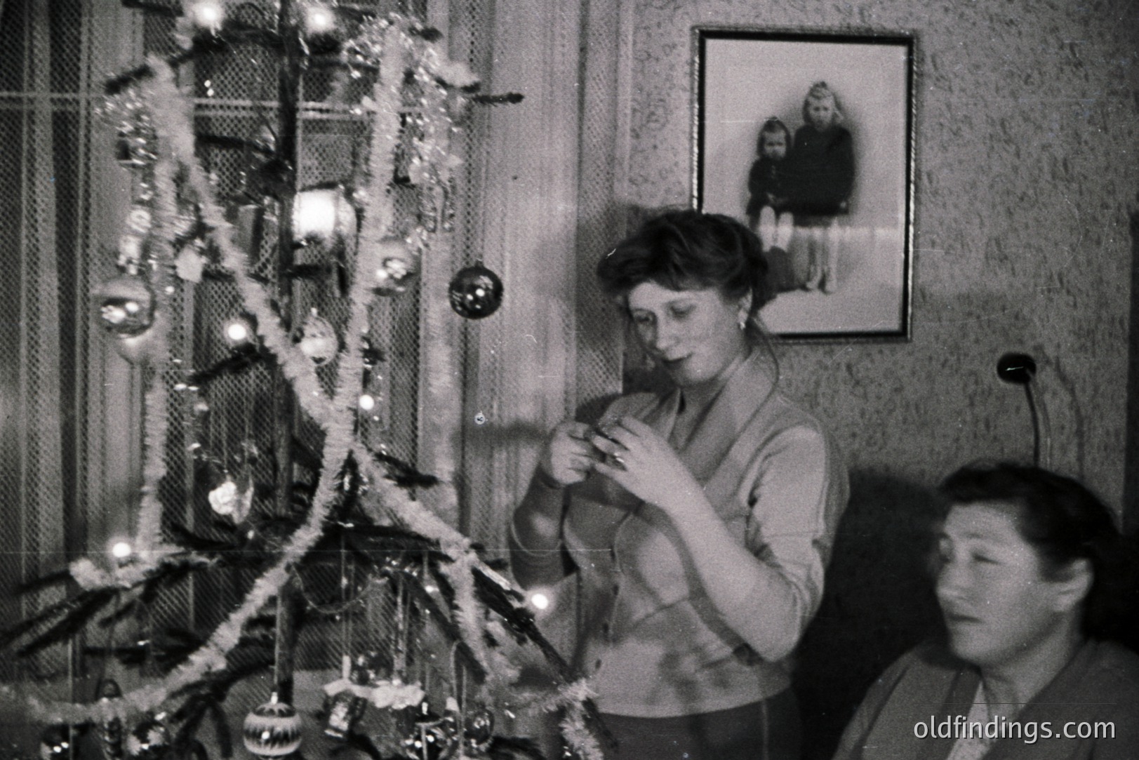 A woman decorates a sparsely-ornamented Christmas tree, possibly in the 1950s or 60s. A framed portrait of two children hangs on the wall. The room exhibits mid-century modern decor with textured wallpaper. Appears to be a home interior.