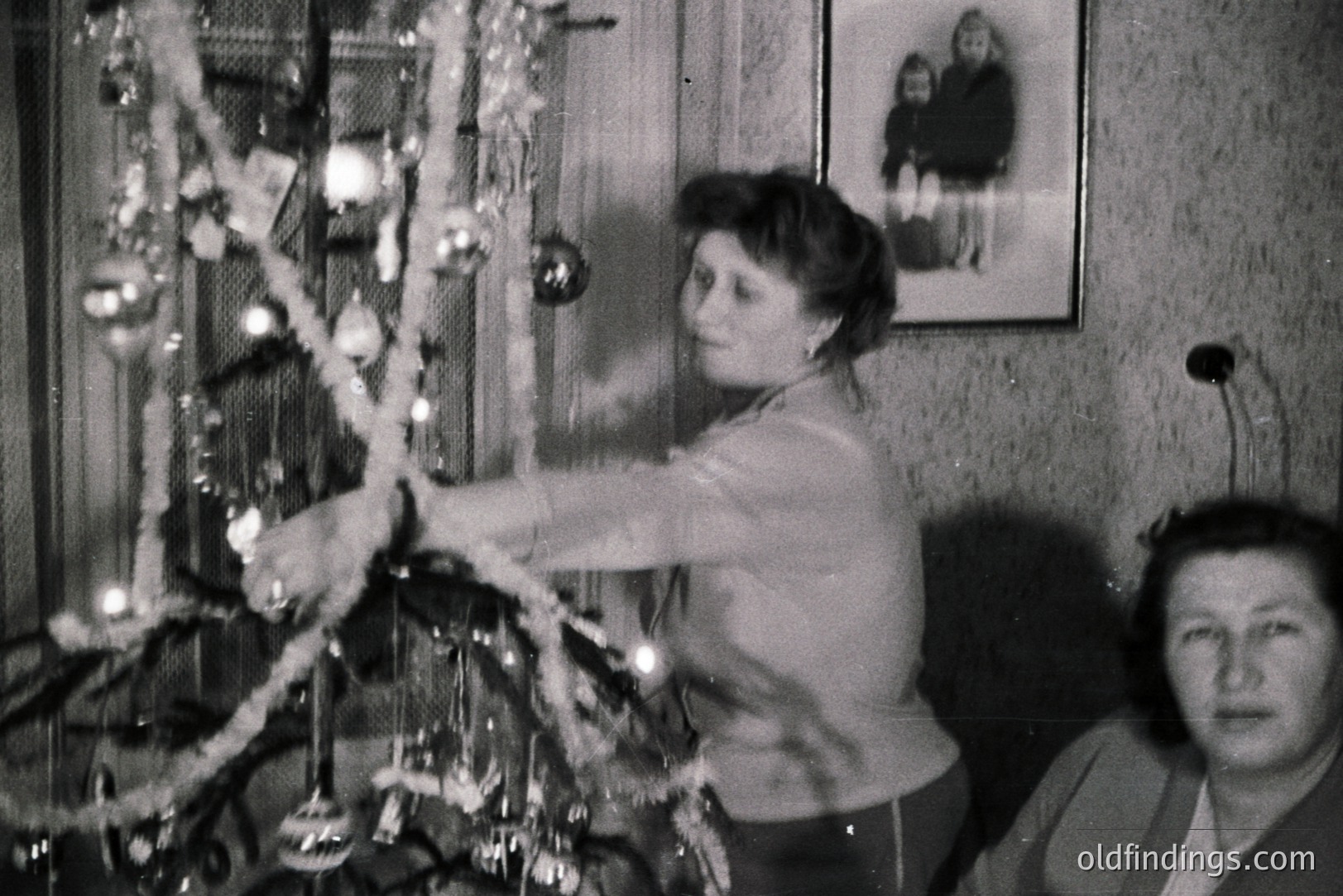 A woman decorates a Christmas tree with garlands, captured in a candid, snapshot-style image. A second woman observes from the side, partially in frame. Likely a family photo from the mid-20th century. Portrait hangs on textured wallpaper. Nostalgic holiday scene.