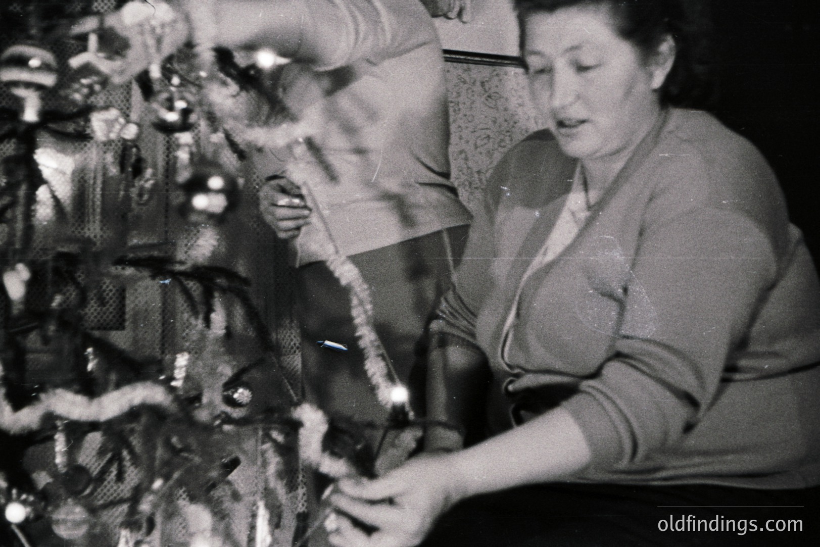 A woman decorates a Christmas tree with string lights, captured in a candid moment. The tree features glass ornaments. Interior shows textured wallpaper and a glimpse of a second person. Likely a mid-century domestic scene, possibly 1950s-1970s. Evokes nostalgia and family traditions.