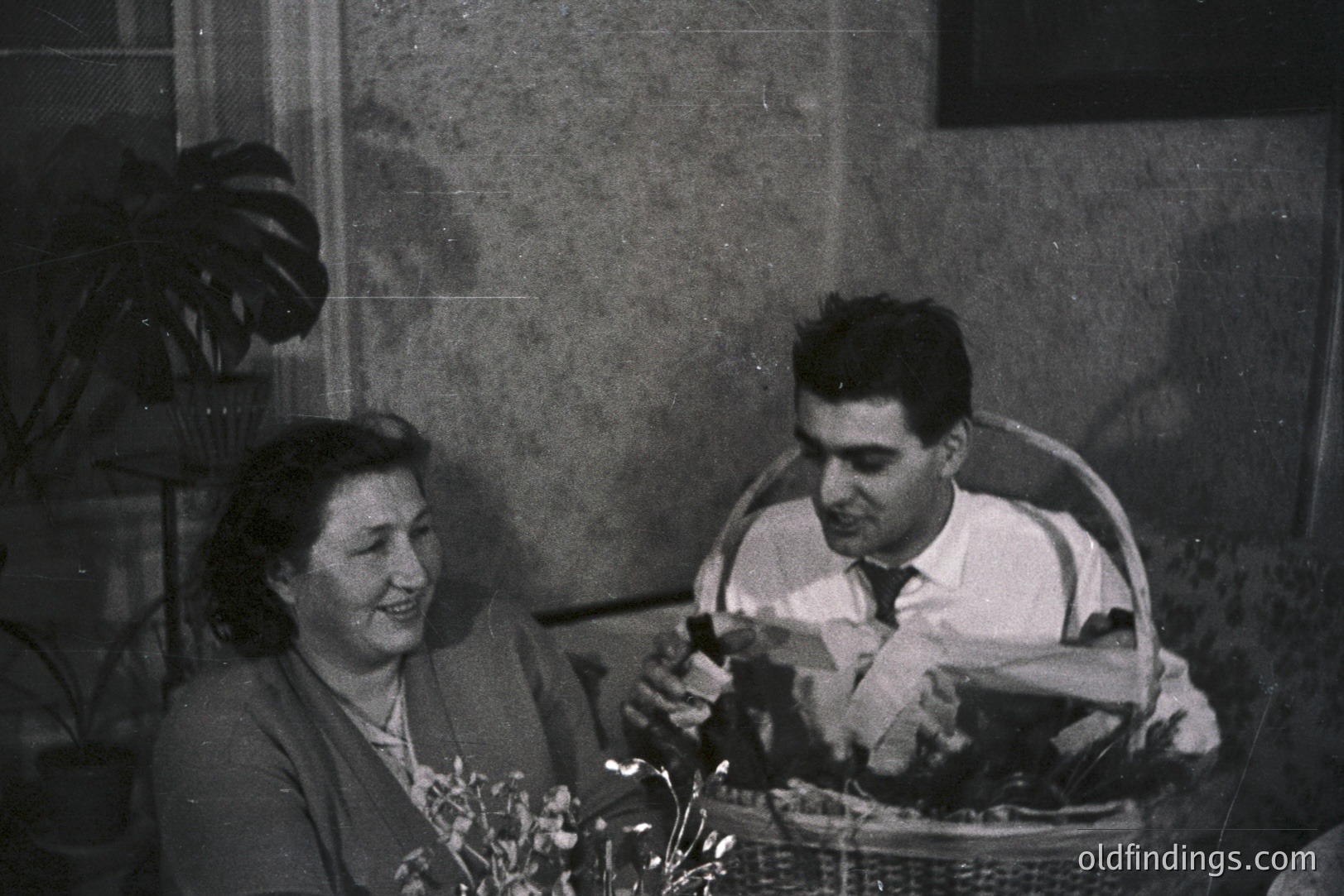 A seated couple shares a moment of joy indoors. The woman, wearing a sweater, smiles while the man, in a suit and tie, presents a gift wrapped in patterned paper. A wicker basket overflowing with foliage sits between them. Likely a family gathering, possibly 1950s.