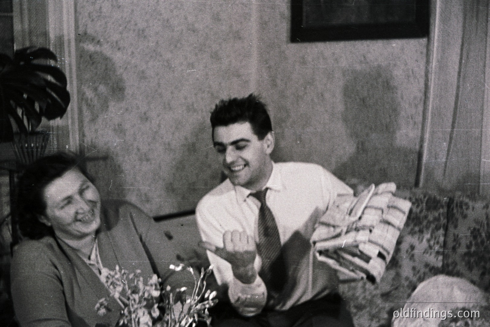 A seated woman smiles while a man, smartly dressed in a collared shirt and tie, playfully presents a wrapped gift. Likely a family portrait, possibly a celebration. Interior scene with foliage and mid-century furnishings suggests the 1950s-1960s. Domestic scene holds potential for nostalgia marketing.