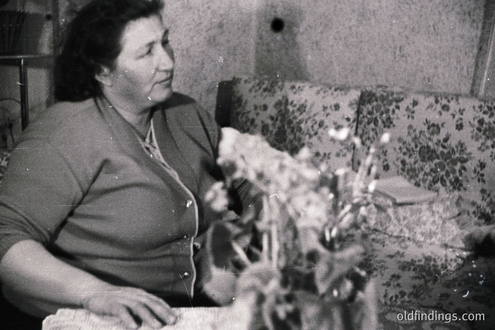 A woman seated at a table with floral arrangement. She wears a button-down shirt with a collar. Interior setting with floral patterned couch visible in background; slight damage/aging on image. Likely a domestic portrait from the mid-20th century.