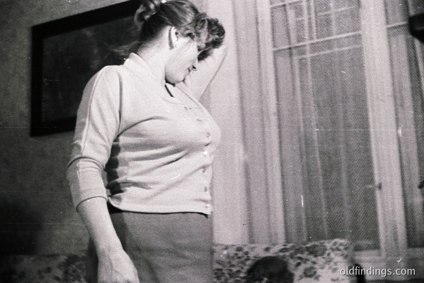 A young woman stands near a window, gently touching her forehead. She wears a button-down sweater and skirt, indicative of 1960s-70s style. A framed picture hangs on the wall, partially visible. Floral patterned armchair in the lower right. Likely a candid, personal snapshot.