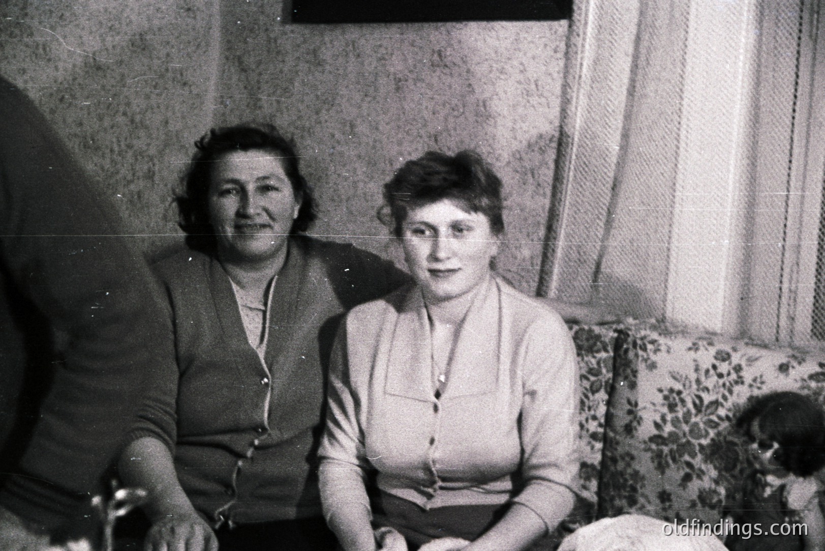 Two women seated on a floral patterned sofa. The woman on the left wears a cardigan over a high-necked blouse, while the woman on the right has a shorter hairstyle and a button-down shirt. A small child is partially visible on the far right. Likely a domestic snapshot, possibly 1960s-1970s.