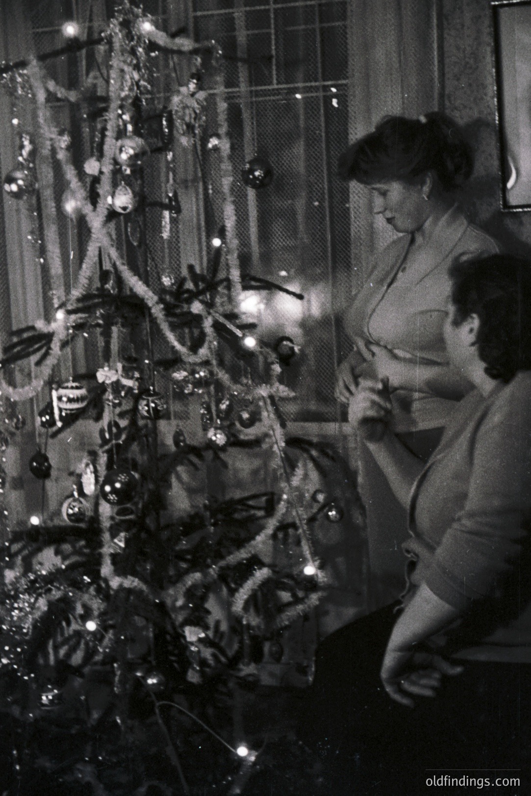 Two women decorate a tinsel-laden Christmas tree indoors, likely in the 1950s or 60s. Natural light and decorative lights illuminate the scene. The tree’s sparse branches are adorned with glass ornaments and tinsel. A vintage domestic moment, evocative of mid-century holiday traditions.
