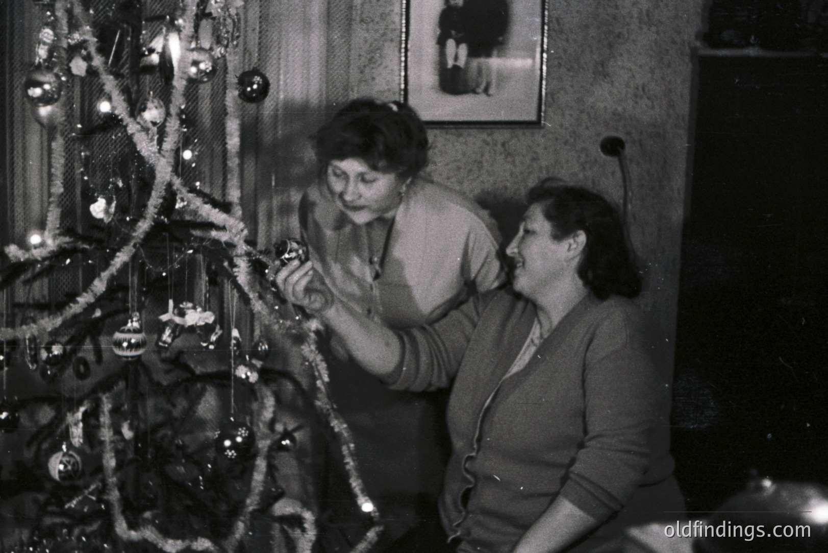 Two women decorate a Christmas tree in a cozy, modestly furnished room. A framed portrait hangs above the tree, suggesting a family connection. Likely a domestic snapshot from the mid-20th century, capturing a quiet moment of holiday tradition.