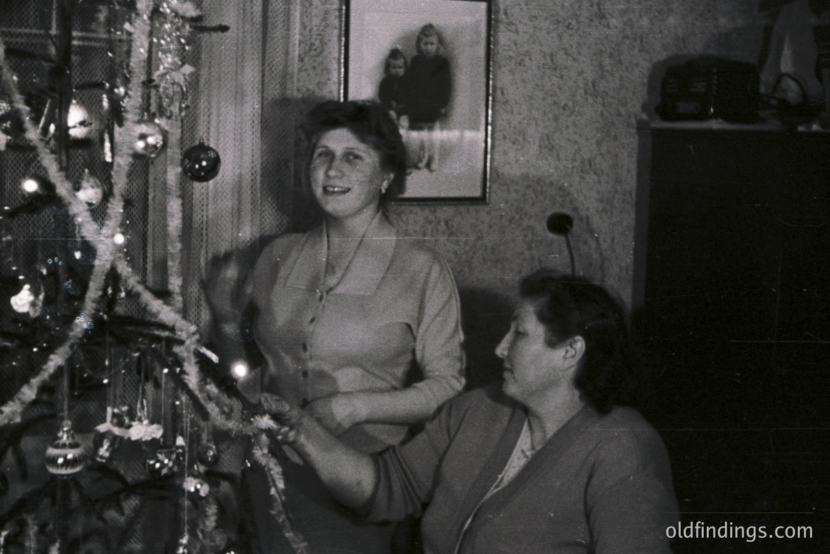 Two women decorate a Christmas tree in a modestly furnished room. A framed portrait hangs on the wall. Likely a 1950s or 60s family snapshot, exhibiting mid-century home décor. The scene captures a domestic moment of holiday preparation.