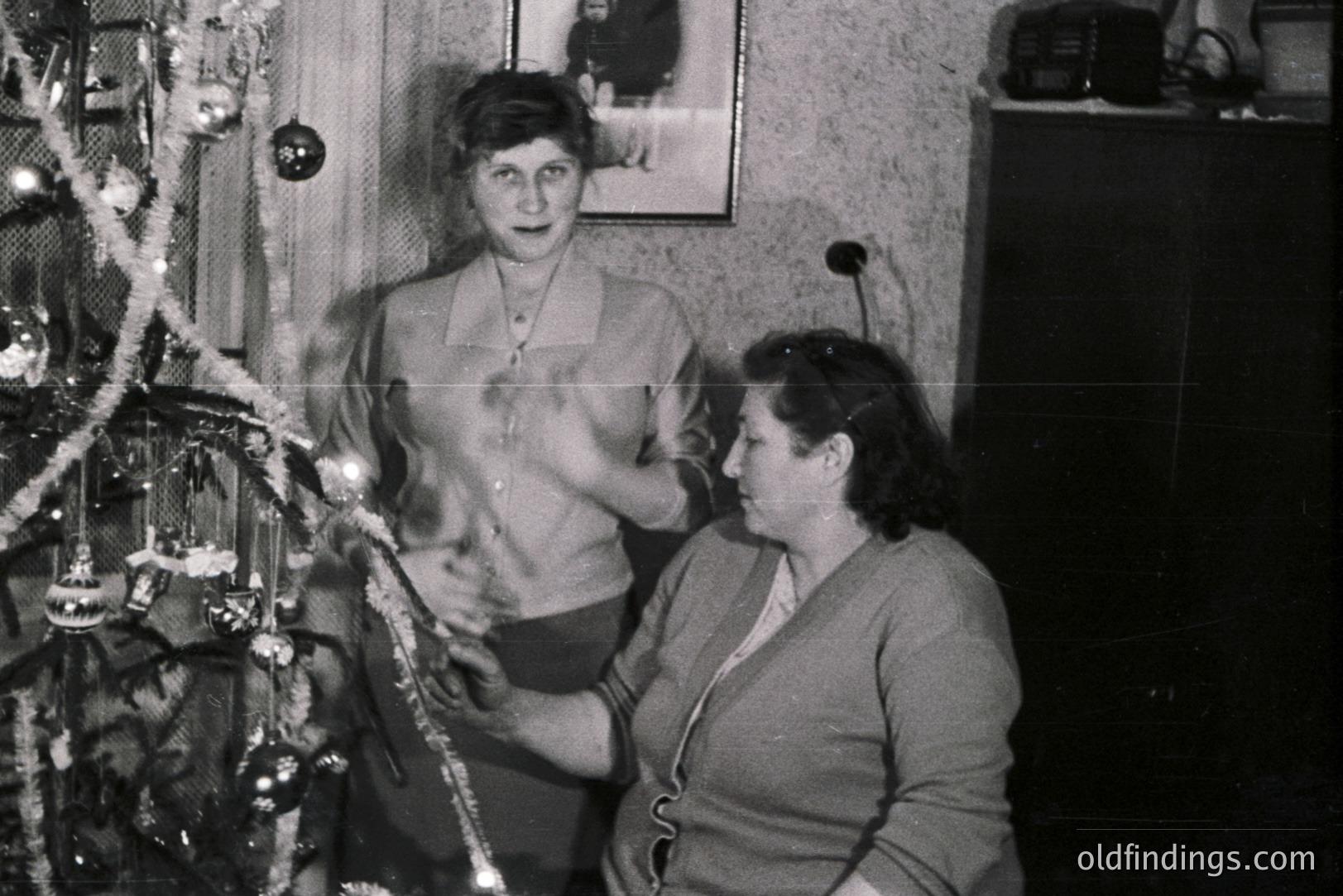 A black and white snapshot captures two women decorating a Christmas tree indoors. One stands, adjusting a garland; the other sits nearby, possibly assisting. The tree is adorned with tinsel and ornaments. A framed portrait hangs on the textured wall. Likely taken in the mid-20th century, potentially the 1950s or 60s.