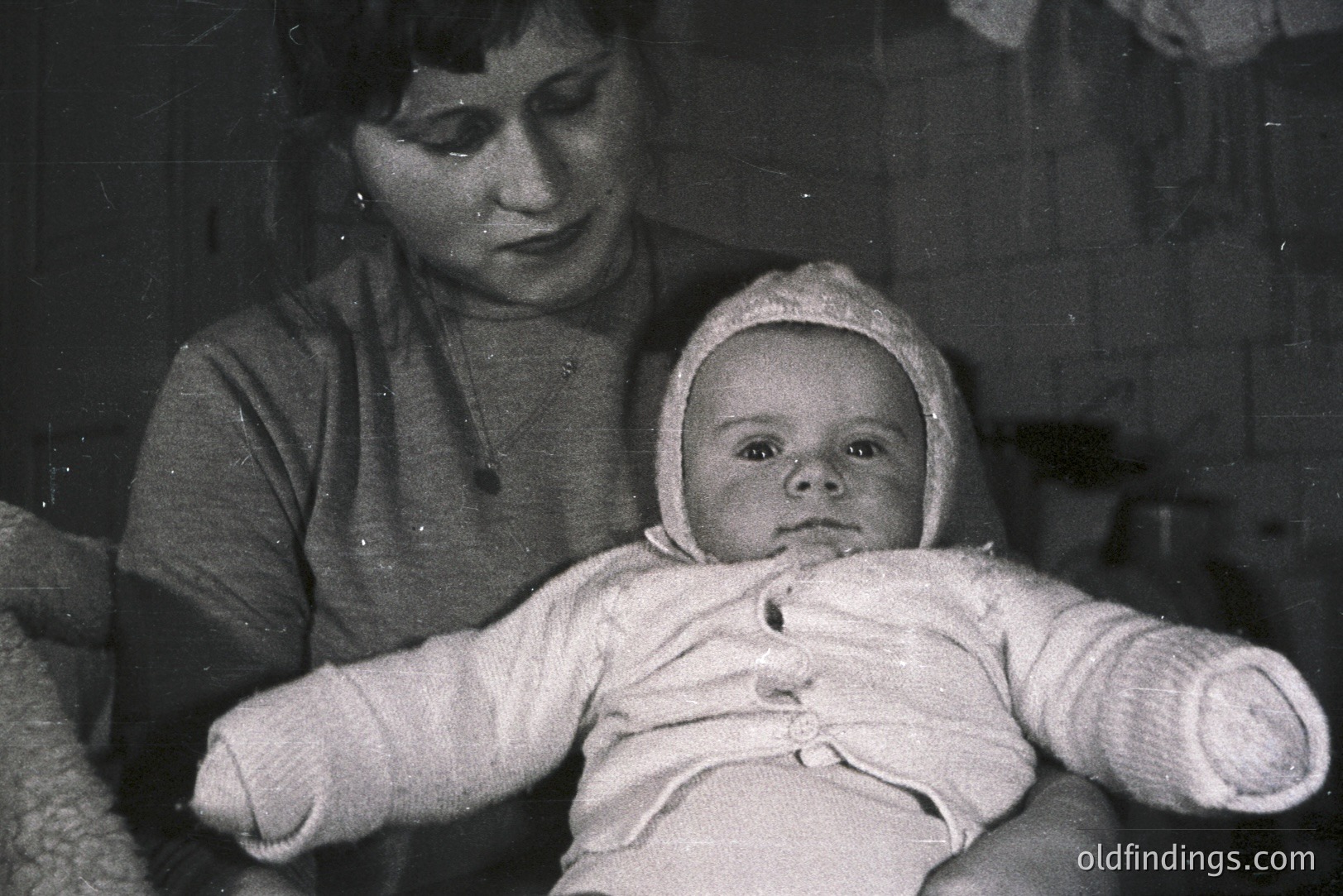 A grayscale portrait features a young woman holding a baby bundled in a bonnet and clothing. The background suggests a simple, indoor setting, possibly a home interior. The image's texture hints at a vintage print, likely from the mid-20th century. The woman wears a simple sweater and a necklace.