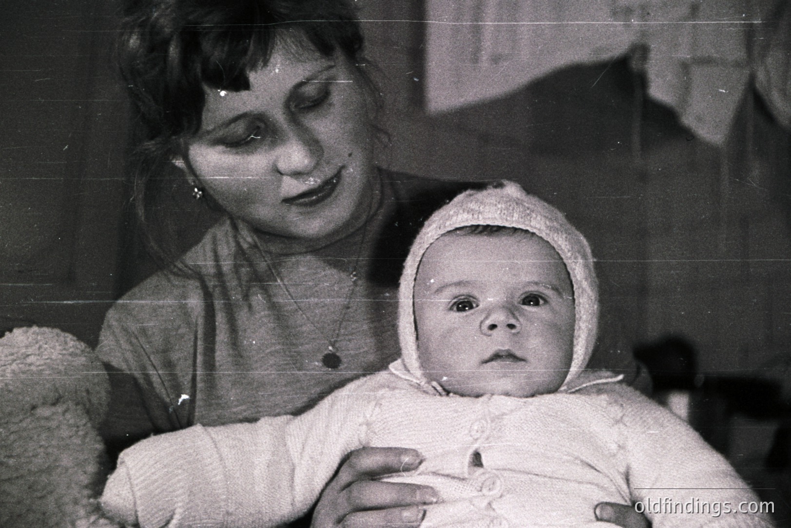 A woman cradles a baby bundled in a heavily textured, knitted bonnet and sweater. Black and white photography suggests a mid-20th century domestic setting. The woman wears a simple sweater and a delicate necklace. A soft, fuzzy blanket is visible in the lower left corner. This could be a family portrait or informal snapshot.