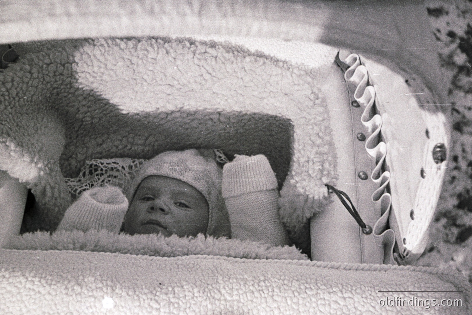 Black and white photograph of an infant nestled within a heavily padded, ornate baby carriage. The baby wears a knitted bonnet and mittens, partially obscured by delicate lace. Carriage details suggest a vintage design, possibly 1940s-1960s. Documenting a moment of early childhood.