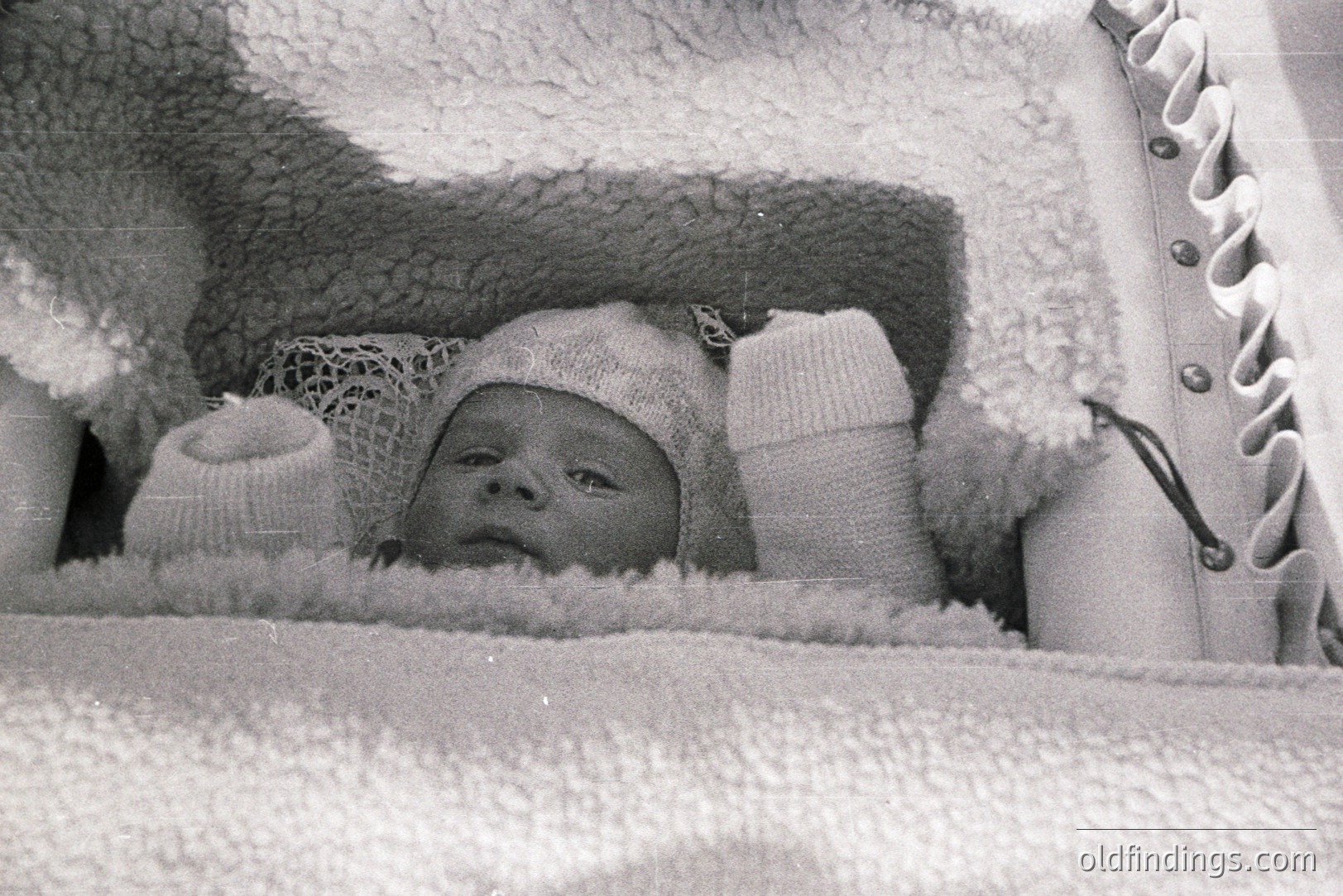 Infant securely nestled in a cushioned carrier with lace-trimmed bonnet and mittens. The wicker structure suggests a vintage baby carrier, likely from the 1950s-1970s. The soft focus and black-and-white aesthetic convey a nostalgic, intimate moment.