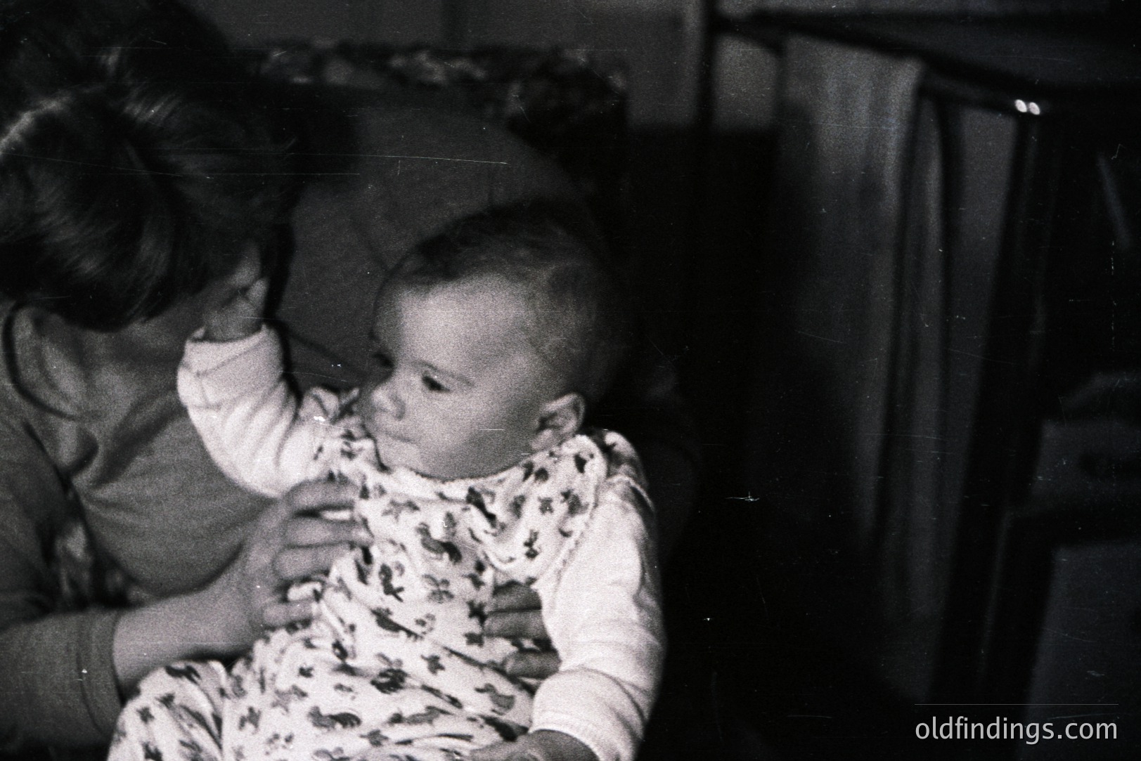 A black and white photo captures a baby held by an adult. The child wears a patterned onesie, gazing directly at the camera. Soft lighting and shallow depth of field create an intimate, candid portrait, likely a personal snapshot. Appears to be mid-century domestic photography.