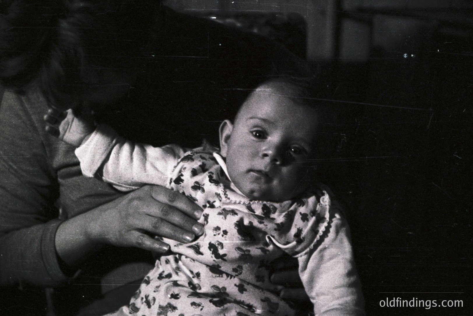 Black and white portrait of a baby held by an adult. The child wears a patterned romper, looking directly at the camera. A hand gently rests on the baby's chest. Likely a family snapshot, circa 1950s-1970s. The image exhibits qualities typical of mid-century photography.