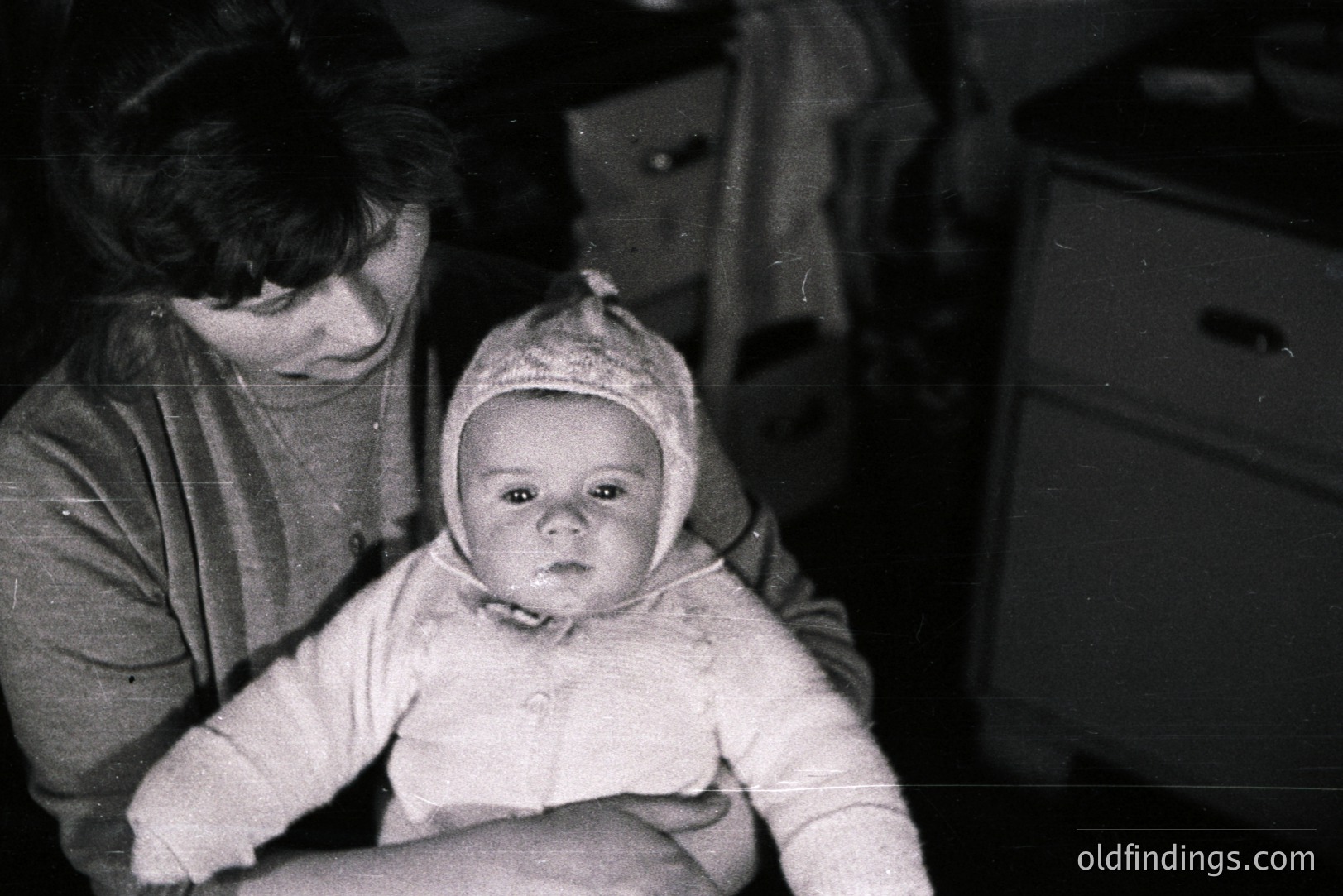 A mother cradles an infant bundled in a soft, textured bonnet and sweater. Interior scene with a dresser and partially visible draped fabric. Likely a domestic snapshot, possibly 1950s-1970s. Focus is on the child; gentle, intimate portrait. Shows subtle signs of age.
