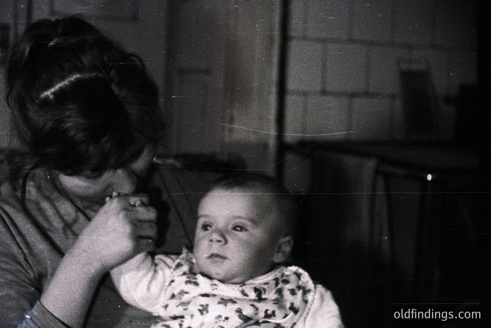 A mother tenderly feeds a baby in a dimly lit interior, likely a kitchen. The baby wears a patterned bib, looking directly at the camera. Photographic style suggests a casual, intimate moment, possibly a snapshot from the 1960s. Background features brickwork and a glimpse of a window. Family portrait.