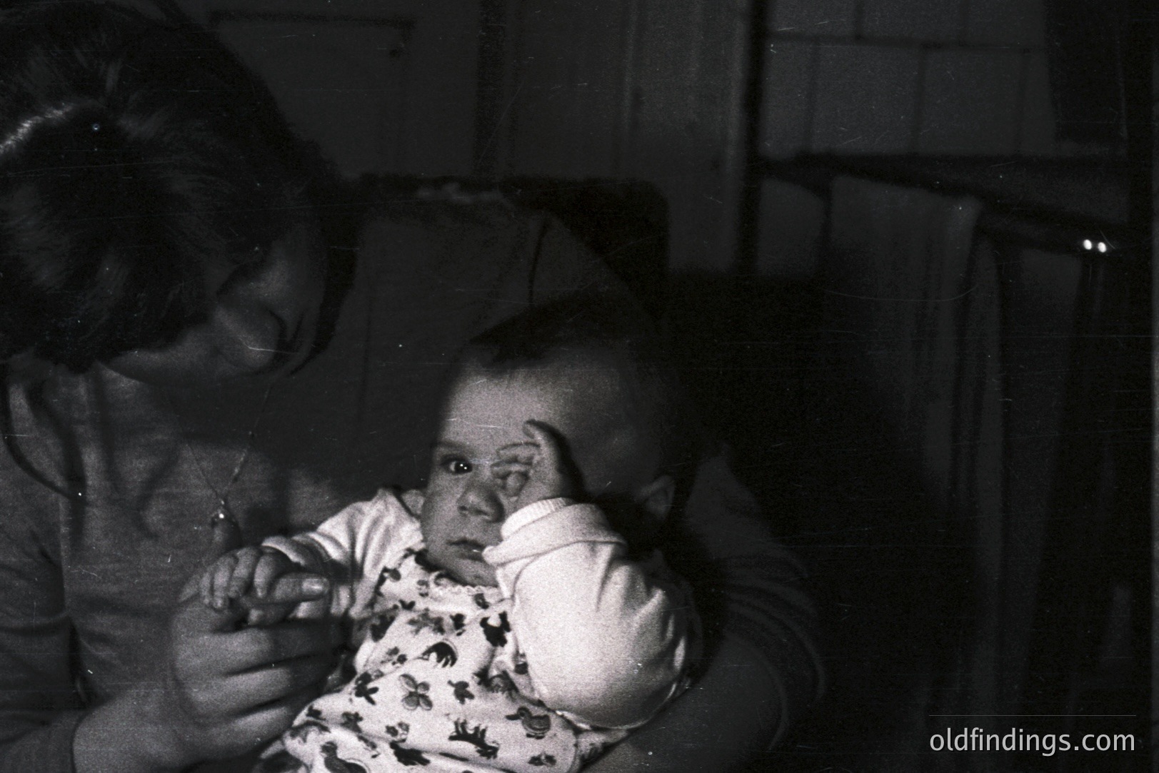 A black and white image captures a young child held by an adult, likely a parent. The child, dressed in a patterned onesie, rests a hand on their head, gazing directly at the camera. The adult’s face is partially visible. Appears to be a personal snapshot, indicative of mid-20th century family photography.