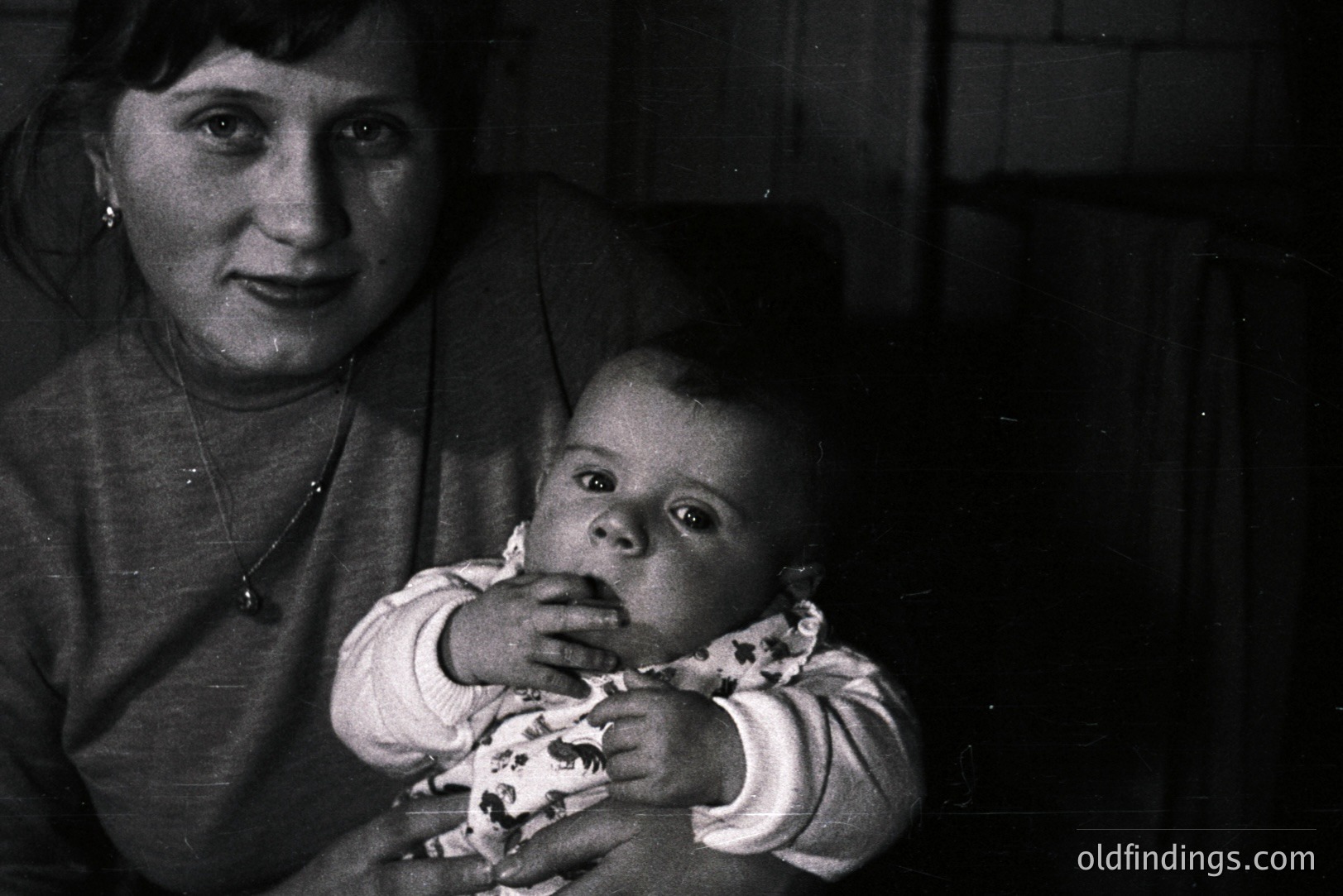 Close-up, monochrome portrait of a woman holding a baby. The woman wears a sweater & necklace; baby in patterned clothing appears curious. Simple, blurred background suggests an interior setting, possibly a home. Likely mid-20th century documentation or personal snapshot. Strong potential for historical research/family archive use.