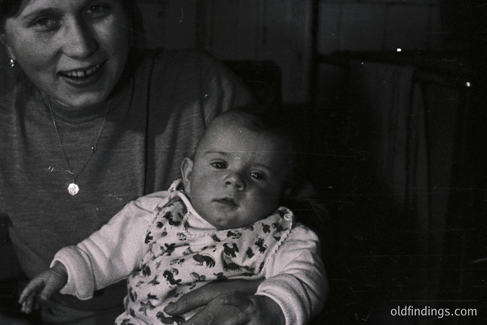 A young mother smiles gently as she holds a baby with wide, observant eyes. The baby wears a patterned bib. Likely a candid family portrait, evidenced by the informal pose and grainy texture. Estimated timeframe: 1950s-1970s. Evokes nostalgia and domestic intimacy.
