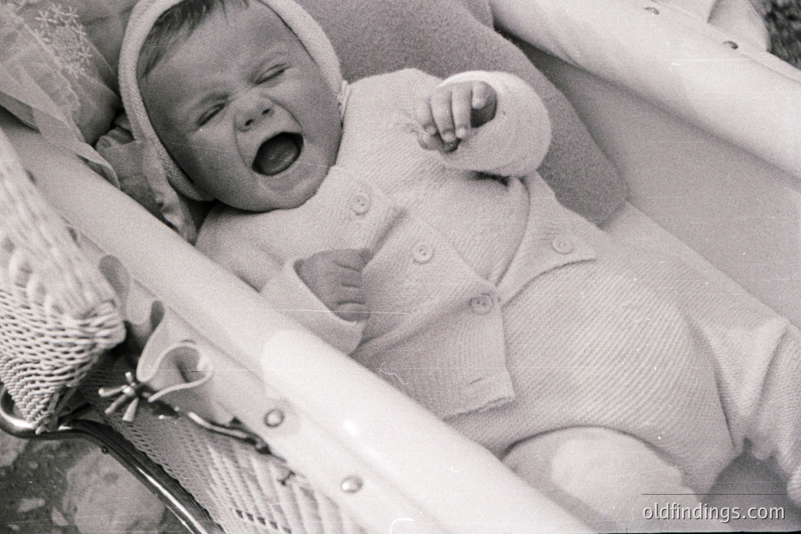 Infant in a vintage wicker travel crib, wearing a knitted bonnet and matching sweater, appears to be crying. Crib details suggest 1960s or 70s design. Image exhibits characteristic grain of film photography. Baby’s pose and attire evoke nostalgic family portraiture.