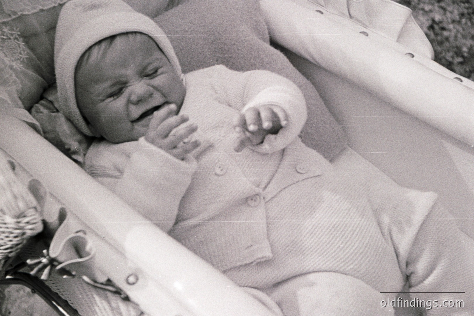 Infant in a close-knit, ribbed sweater and bonnet sits in a wicker carriage, visibly distressed with mouth open and hands raised. Likely a staged portrait, possibly family documentation. The grainy quality suggests a mid-20th century snapshot. High commercial value for vintage baby/family stock.