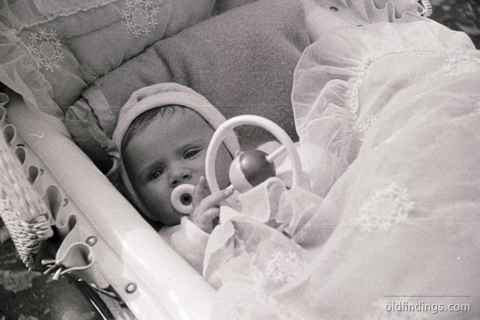 A baby in a wicker cradle, swaddled in elaborate lace and a bonnet, gazes directly at the camera. The cradle’s metal frame is visible, adding an industrial contrast to the delicate textiles. Likely a family portrait, c. 1930s-1950s. Potential value for vintage baby/family studies.