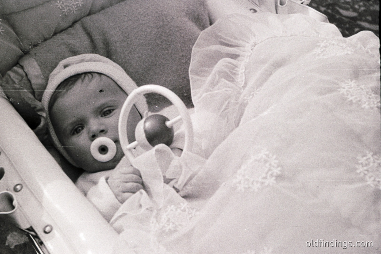 Infant in a vintage carriage, pacifier visible, surrounded by ornate lace and cushioned padding. Likely 1950s-1970s, suggesting a period of domesticity and emphasis on infant care. Carriage design indicative of mid-century styles.