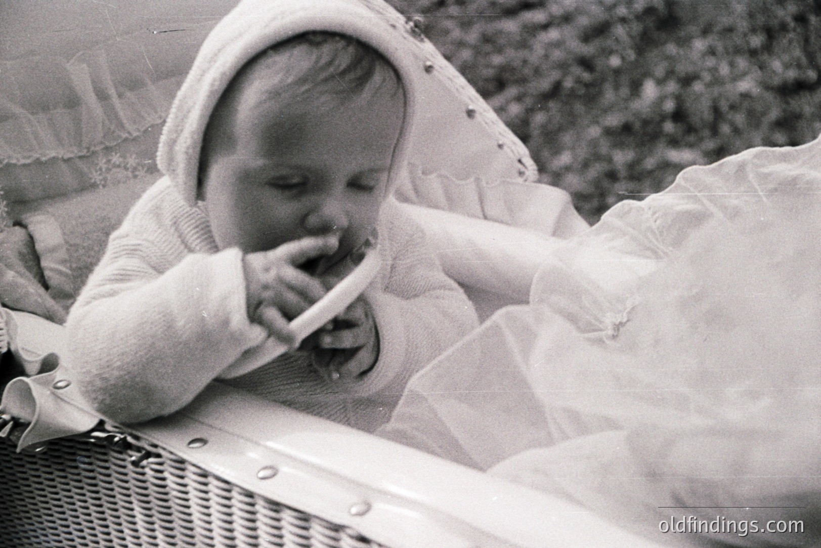 Infant seated in a wicker basket, wearing a bonnet and long-sleeved dress. Appears to be chewing on a teether. Likely a candid family photograph, reflecting mid-century parenting styles. Grainy black & white image suggests a 1950s-1960s origin. Potential for nostalgia-themed design.