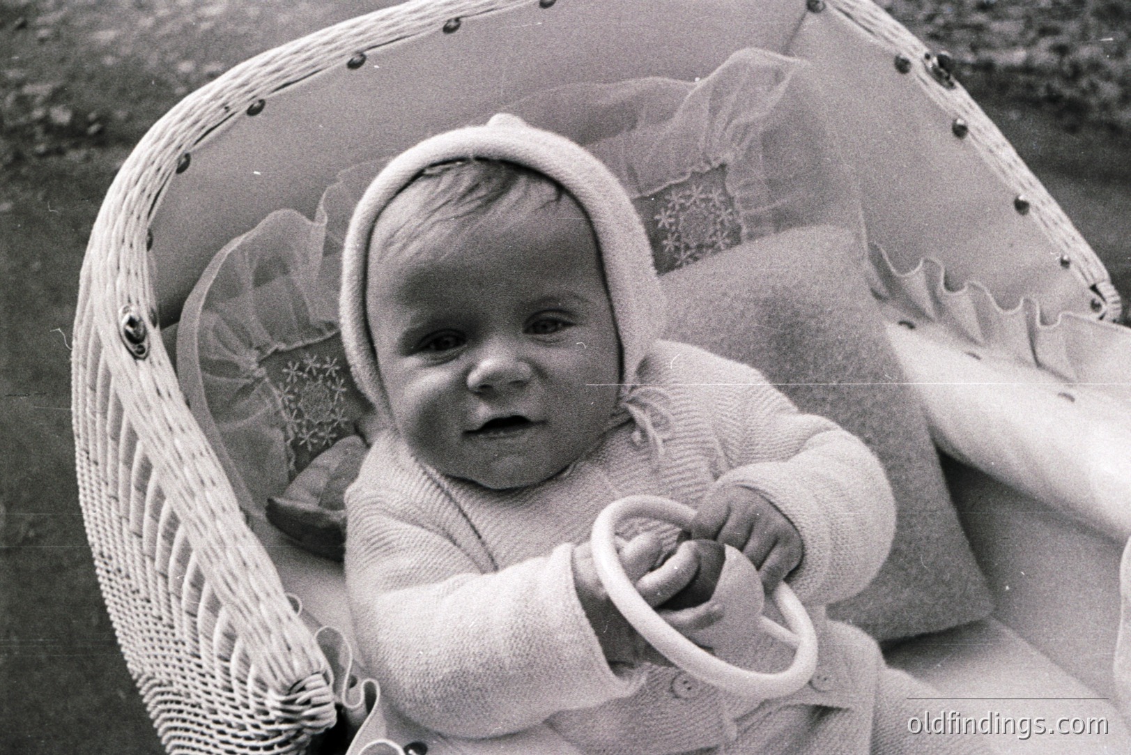 Infant in a wicker bassinet, adorned with a bonnet and knit romper suit. Small hands grip a toy steering wheel. Likely a family portrait, circa 1950s-1960s. Soft lighting, typical of early photographic techniques. The scene conveys domestic tranquility.