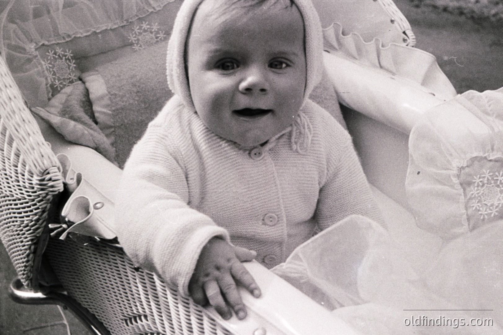 Portrait of a baby, likely around 6-9 months old, wearing a knitted sweater and bonnet, seated in a high chair. Delicate lace detailing on chair and bib. Vintage photography style suggests the 1950s or 1960s. Ideal for nostalgia or family heritage projects.