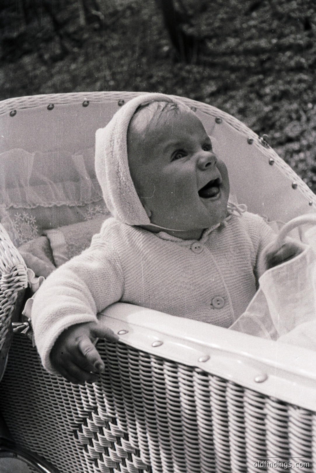 A baby, bundled in a knitted bonnet and sweater, expresses joyful surprise from within a wicker baby carriage. Classic mid-century design suggests a 1950s or 1960s origin. Gentle outdoor lighting creates a soft, nostalgic atmosphere. Ideal for archival, design reference or vintage stock imagery.