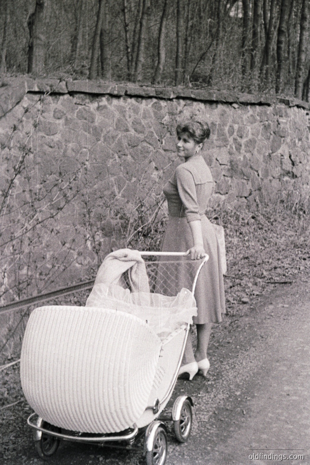 A woman in a mid-length dress and heels stands beside a vintage pram with elaborate metalwork and fabric canopy. The setting appears to be a road or path near a stone wall and foliage. Likely from the 1950s or 60s, showcasing period fashion and baby transport. A strong visual for design or vintage advertising.